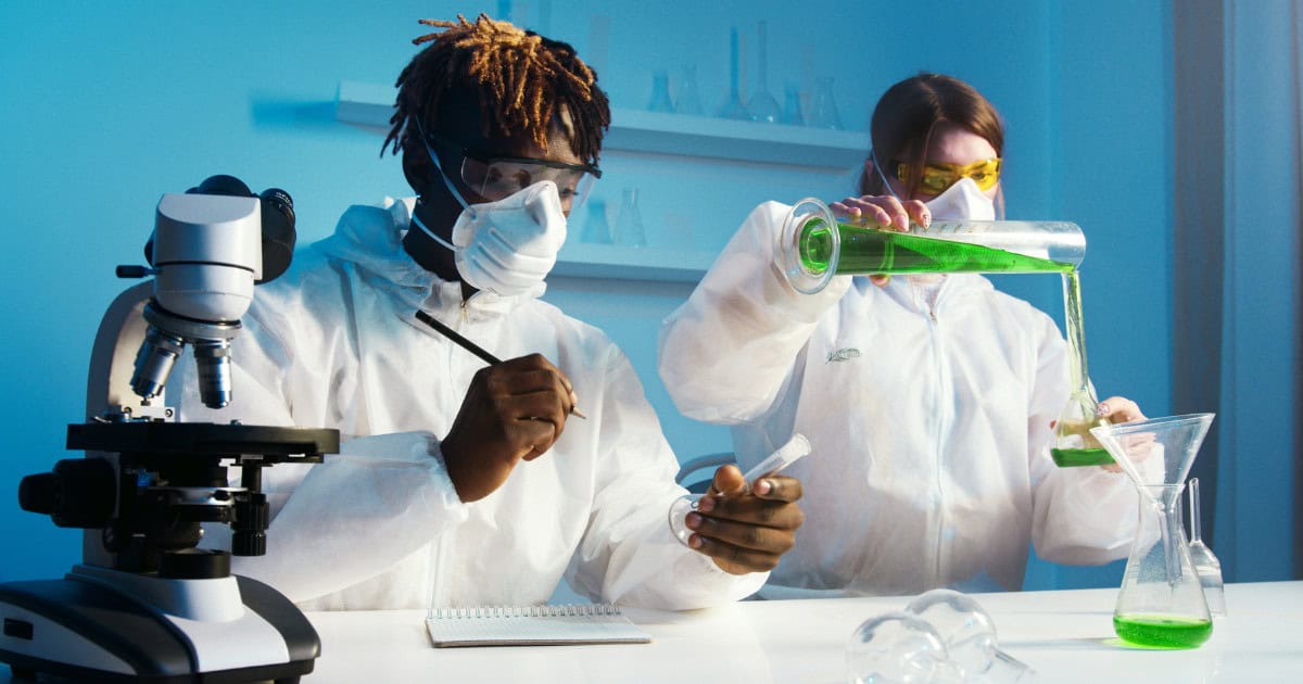 Student in a chemistry lab, wearing goggles, with beakers and test tubes in the foreground, appearing focused on an experiment.