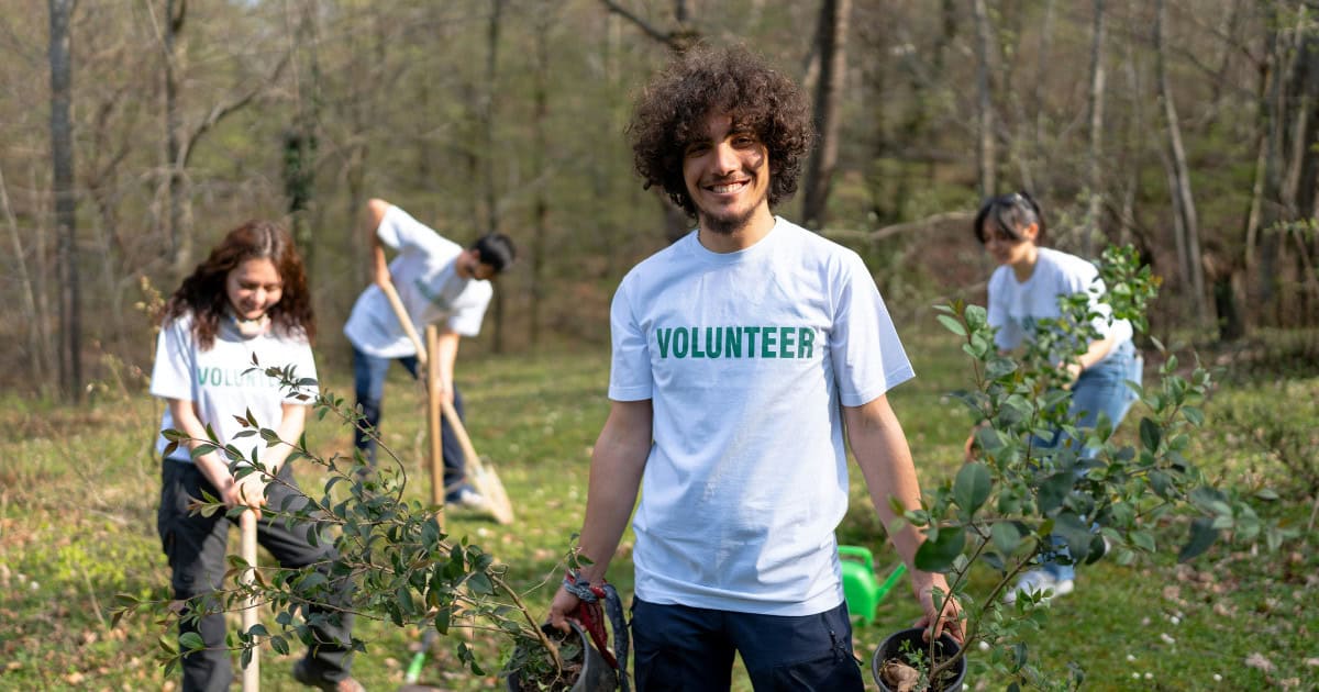 A diverse group of students volunteering at a community event, planting trees, and smiling at the camera.