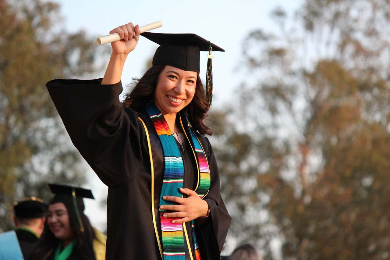 A smiling female graduate in a cap and gown holds up her diploma proudly during a graduation ceremony.