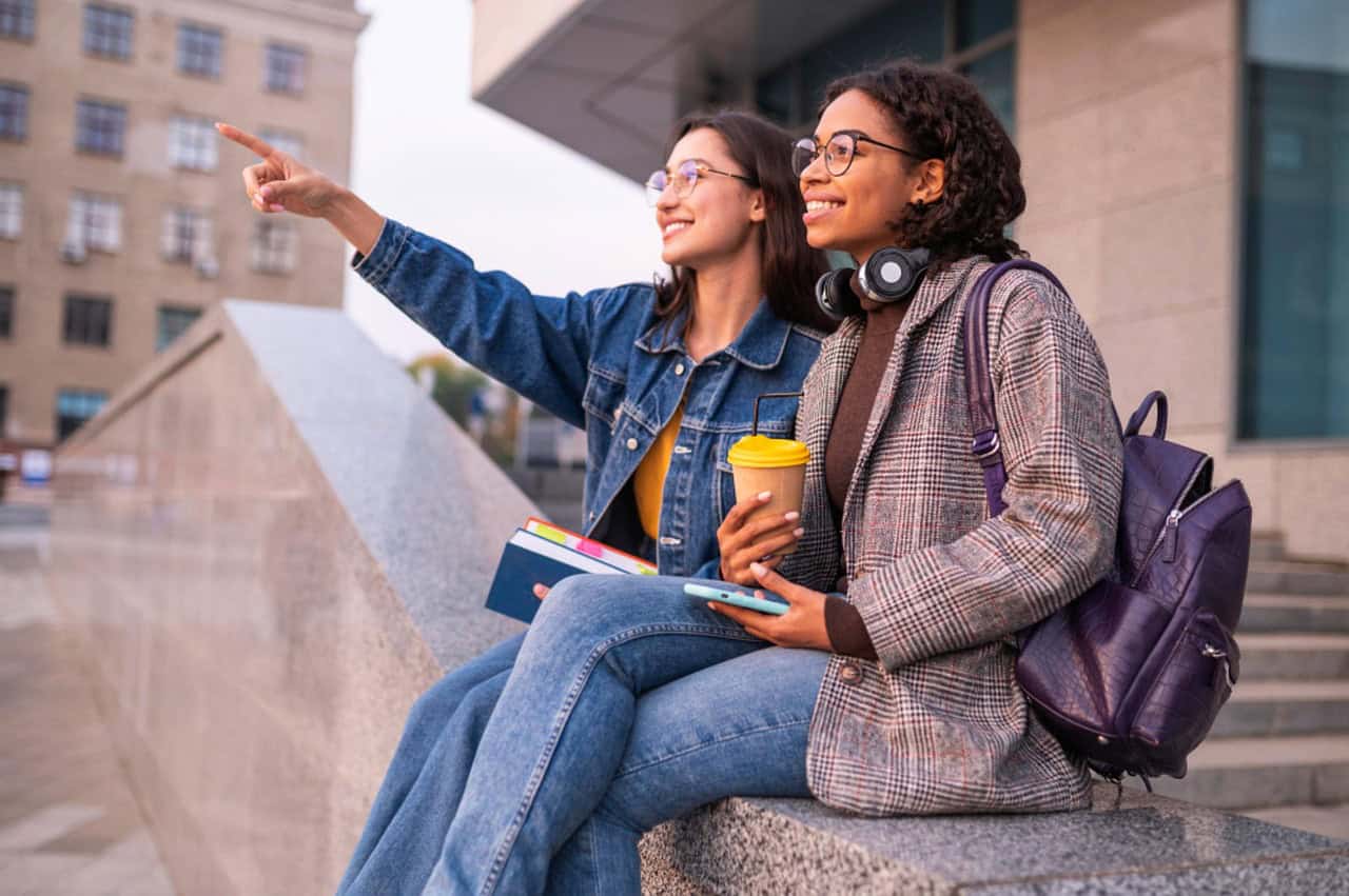 Two college students sitting outside a campus building, as they explore college options.
