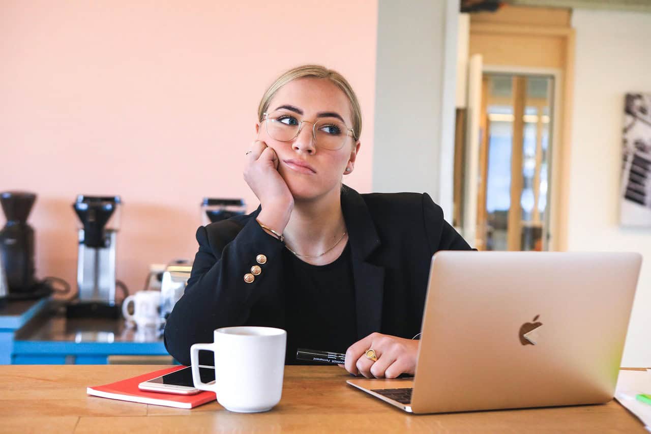 A young woman sits at a desk with a laptop, notebook, and coffee mug with a bored or thoughtful expression.