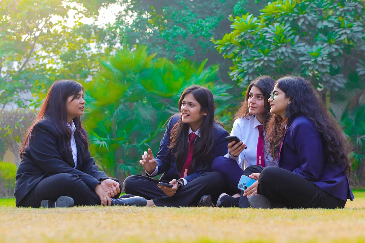 Four college women in formal attire chat and relax on the grass in a sunny outdoor setting.